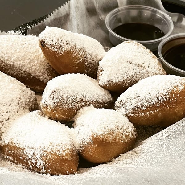 A basket of golden-brown beignets, heavily dusted with powdered sugar, with two small cups of dipping sauce.